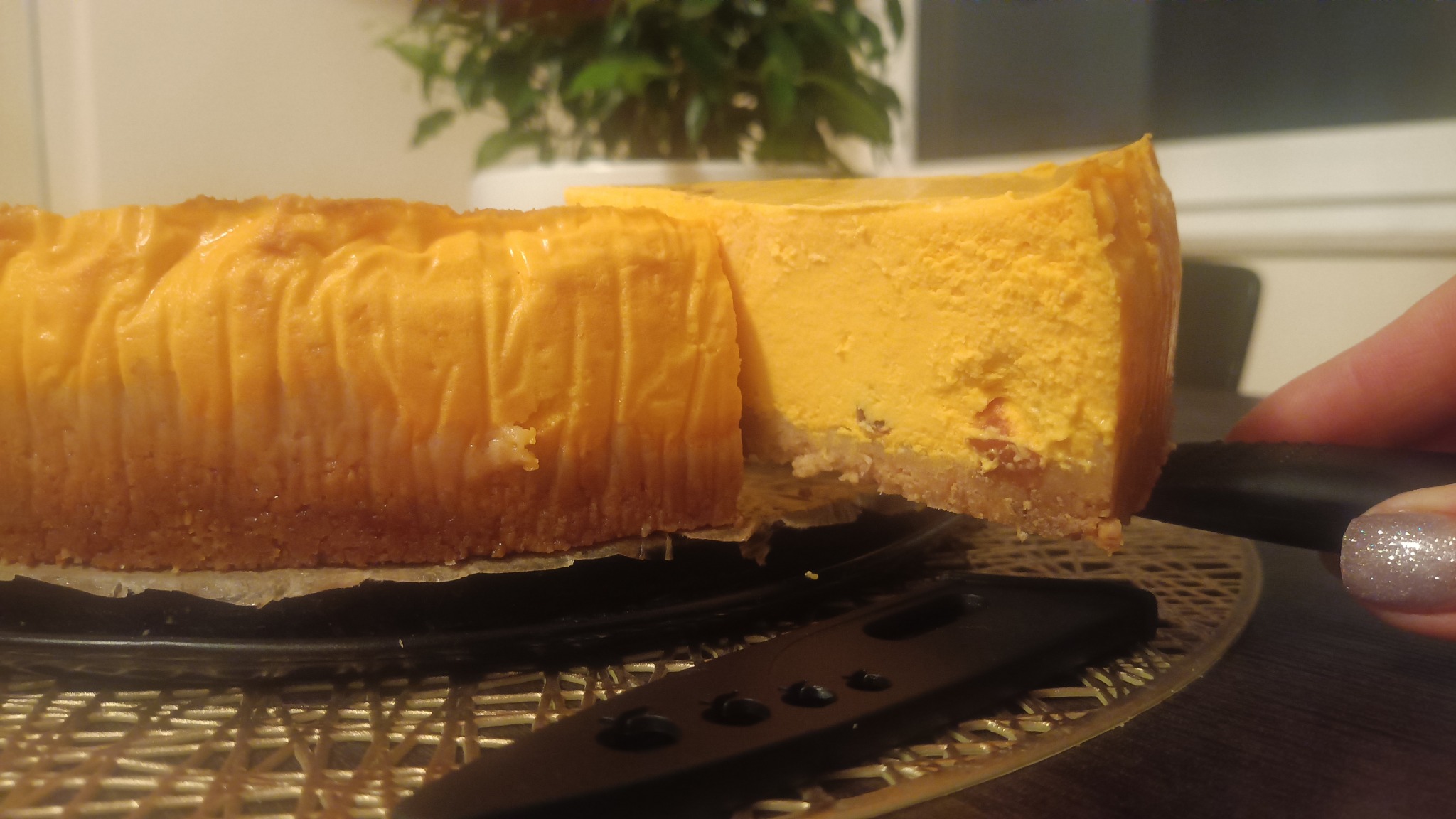 Kawałek pomarańczowego sernika na kruchym spodzie, podawany na czarnym talerzu z ozdobną podkładką, w tle roślina doniczkowa.