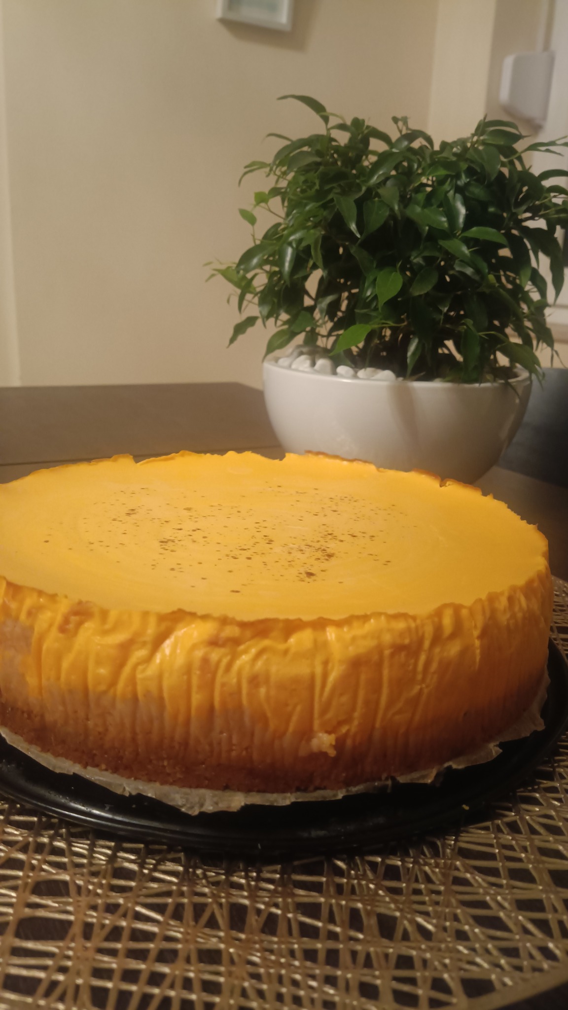 Żółty sernik na czarnym talerzu, z delikatnymi ciemnymi plamkami na wierzchu, ustawiony na plecionym, złotym podkładzie, w tle roślina doniczkowa.