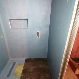 Łukasz szydlik - Wnętrze łazienki w trakcie remontu, ściany pokryte płytami kartonowo-gipsowymi, widoczny otwór na instalację, fragment podłogi z częściowo ułożonymi płytami.