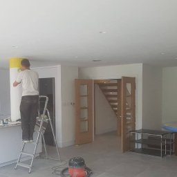 Łukasz szydlik - Mężczyzna na drabinie maluje kolumnę na żółto w nowoczesnej kuchni z wyspą i wiszącymi lampami, widoczne zabezpieczone meble i odkurzacz.