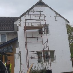 Łukasz szydlik - Dom w trakcie ocieplania styropianem, widoczne rusztowanie, fragment samochodu z otwartą klapą bagażnika i pracownik w kamizelce odblaskowej.