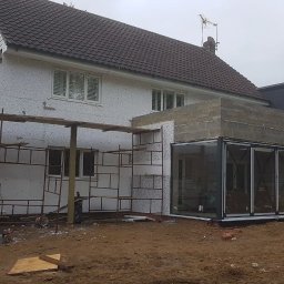 Łukasz szydlik - Dom jednorodzinny w trakcie rozbudowy, widoczne rusztowania i nowa część z dużymi szklanymi drzwiami przesuwnymi, ściany w trakcie ocieplania styropianem.