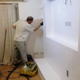 Łukasz szydlik - Mężczyzna w trakcie wygładzania ściany z płyty karton gipsowej, widoczna konstrukcja z wnękami, szpachla nałożona na ścianę, podłoga zabezpieczona folią, odkurzacz przemysłowy obok.