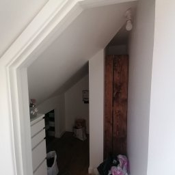 Łukasz szydlik - Pomalowane na biało wejście do poddasza ze skosami, widoczne białe listwy dekoracyjne, surowa żarówka i drewniane drzwi w głębi.