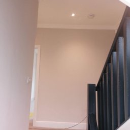 Łukasz szydlik - Jasne wnętrze po odświeżeniu: świeżo pomalowane ściany w odcieniach beżu i szarości, zabezpieczona folią podłoga, widoczne detale sztukaterii sufitowej i czarna balustrada schodów.