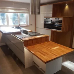 Nowoczesna kuchnia z wyspą, drewnianym blatem i zabudowanymi piekarnikami, oświetlona naturalnym światłem z okna.