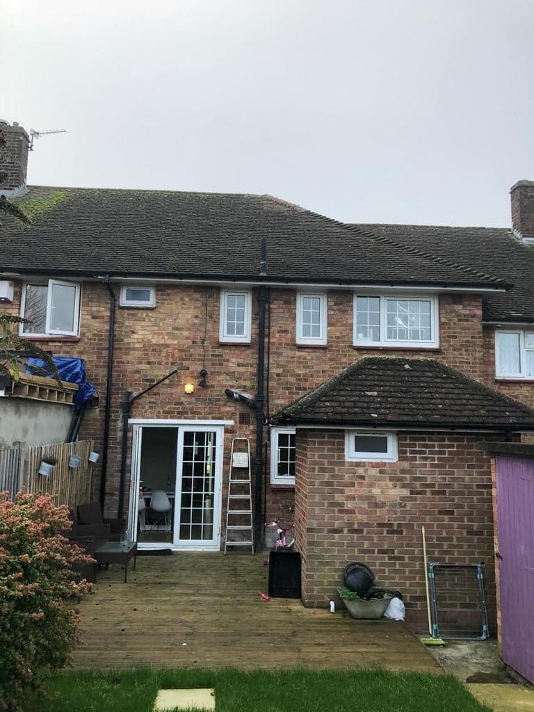 Widok z ogrodu na dwukondygnacyjny dom z cegły z drewnianym tarasem, gdzie widać otwarte białe drzwi balkonowe, drabinę opartą o ścianę i meble ogrodowe.