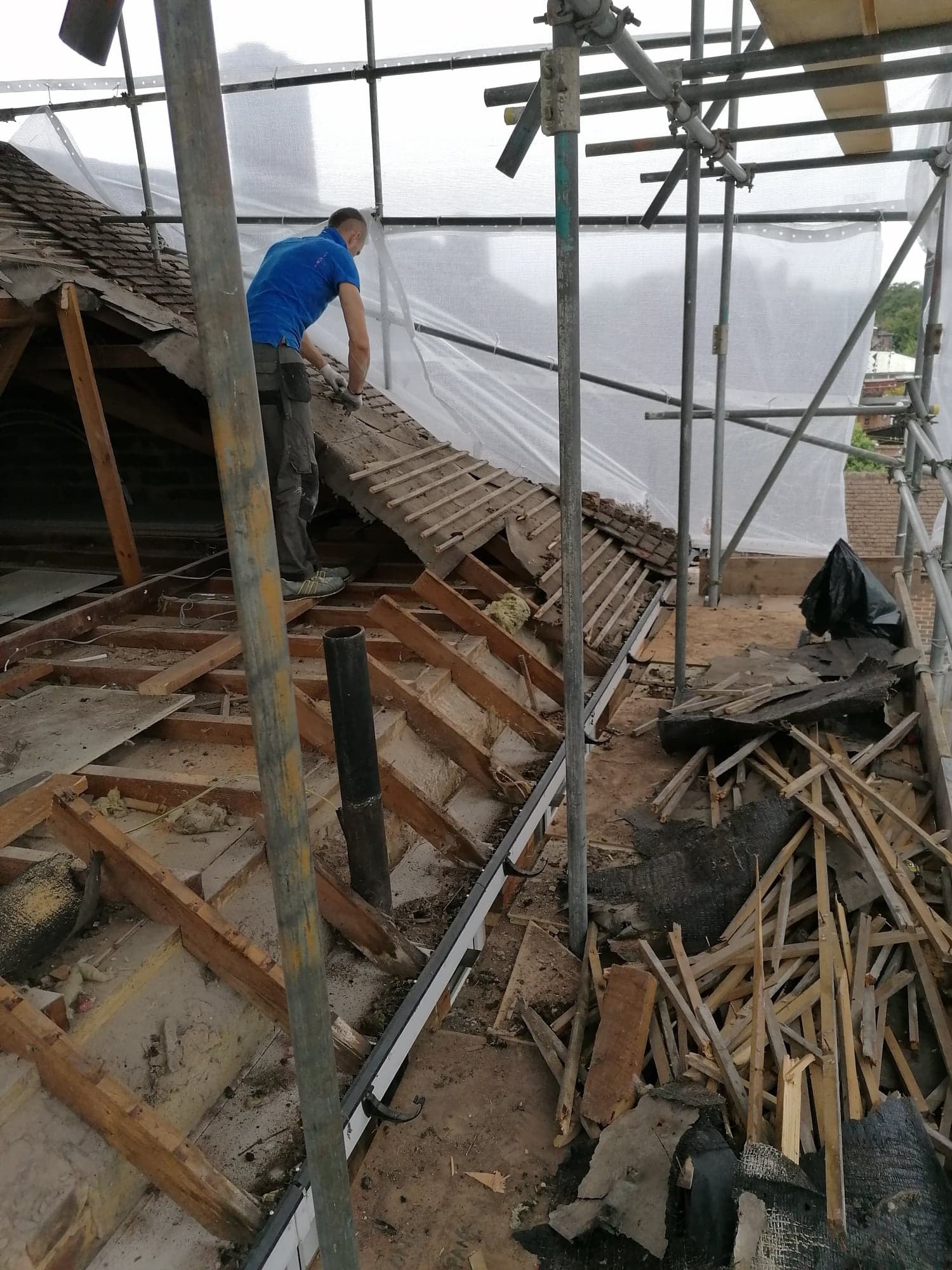 Pracownik w niebieskiej koszulce na rusztowaniu demontuje starą dachówkę z drewnianej konstrukcji dachu, widoczne fragmenty izolacji i gruzu.
