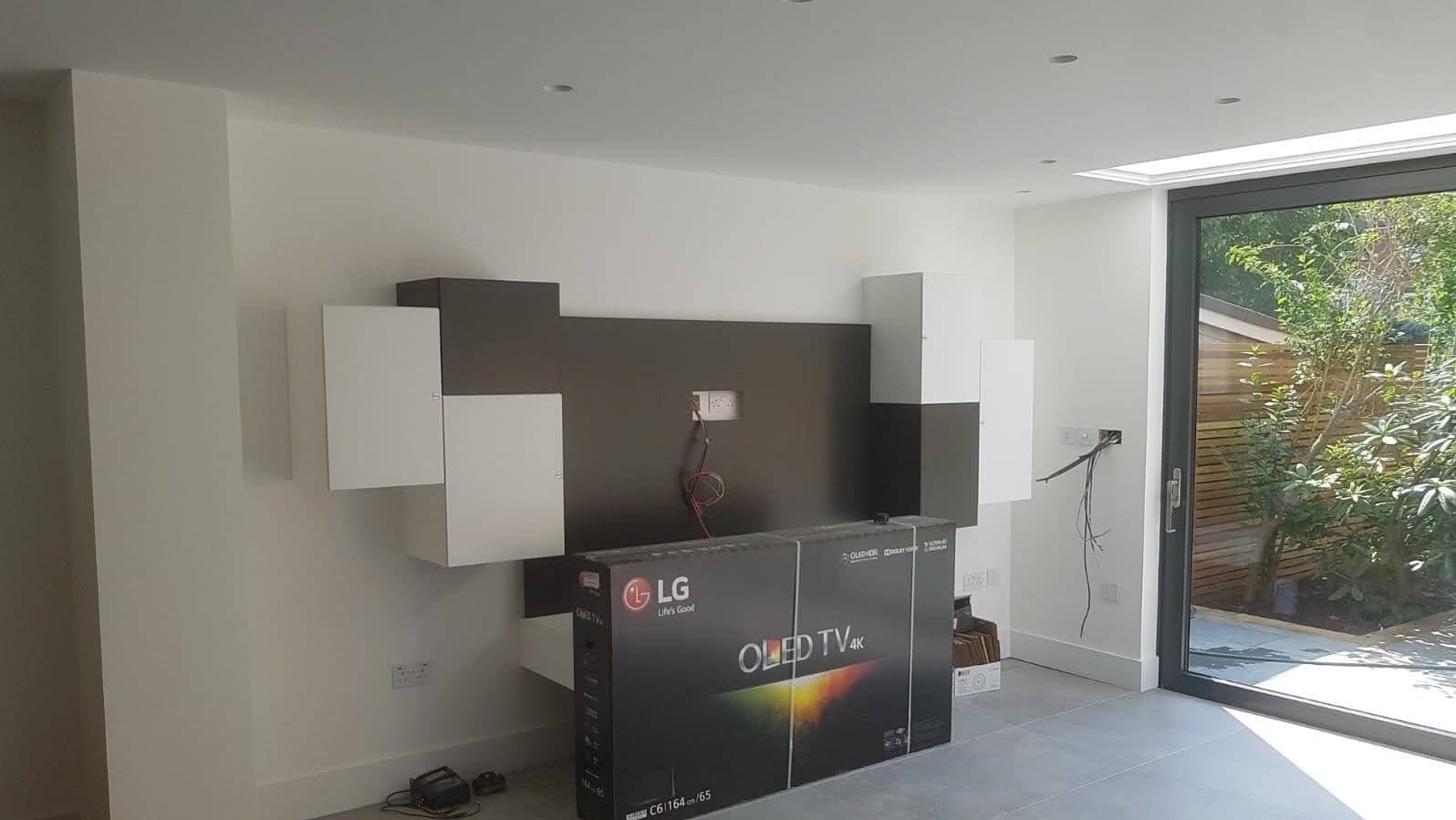 Świeżo wyremontowany salon z nowoczesną zabudową meblową w kolorach bieli i szarości, z przygotowanym miejscem na telewizor, który leży jeszcze w kartonie, widoczne wystające kable i gniazdka...