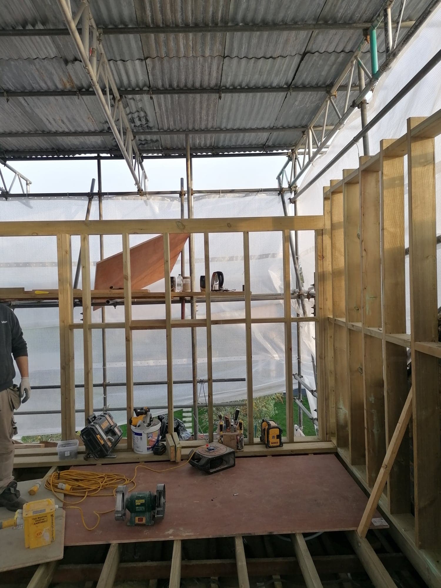 Konstrukcja szkieletowa drewniana w trakcie budowy, widoczne rusztowania, narzędzia i materiały budowlane na podłodze, częściowo osłonięta folią ochronną.