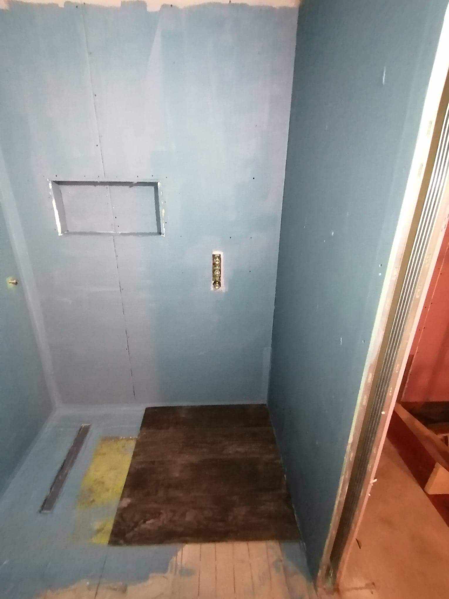 Wnętrze łazienki w trakcie remontu, ściany pokryte płytami kartonowo-gipsowymi, widoczny otwór na instalację, fragment podłogi z częściowo ułożonymi płytami.