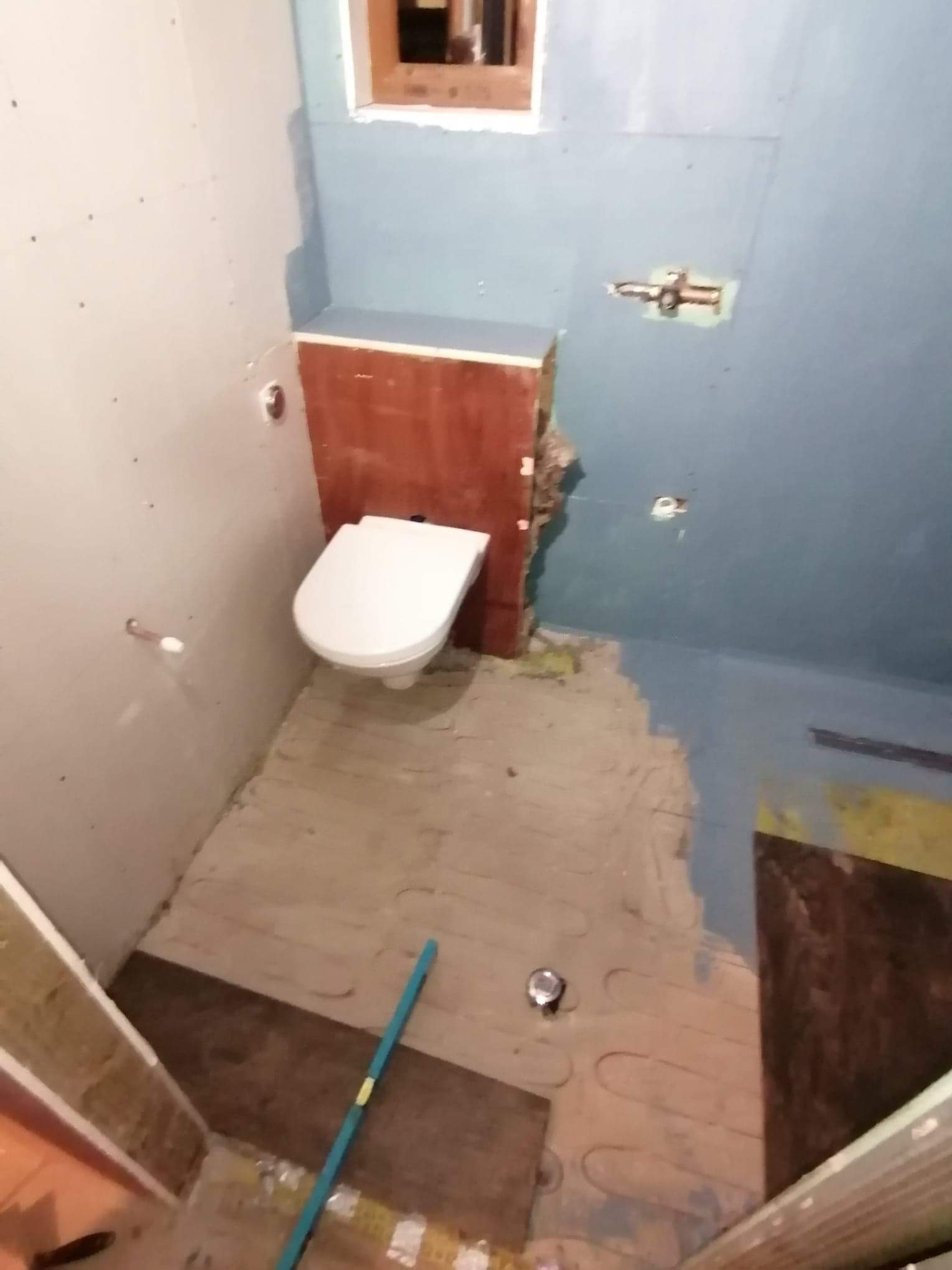 Łazienka w trakcie prac: widoczny zamontowany sedes, surowe ściany z płyt kartonowo-gipsowych i fragmentami pomalowane na niebiesko, podłoga z ogrzewaniem podłogowym przed położeniem płytek.
