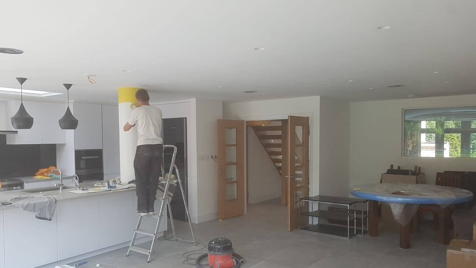 Mężczyzna na drabinie maluje kolumnę na żółto w nowoczesnej kuchni z wyspą i wiszącymi lampami, widoczne zabezpieczone meble i odkurzacz.