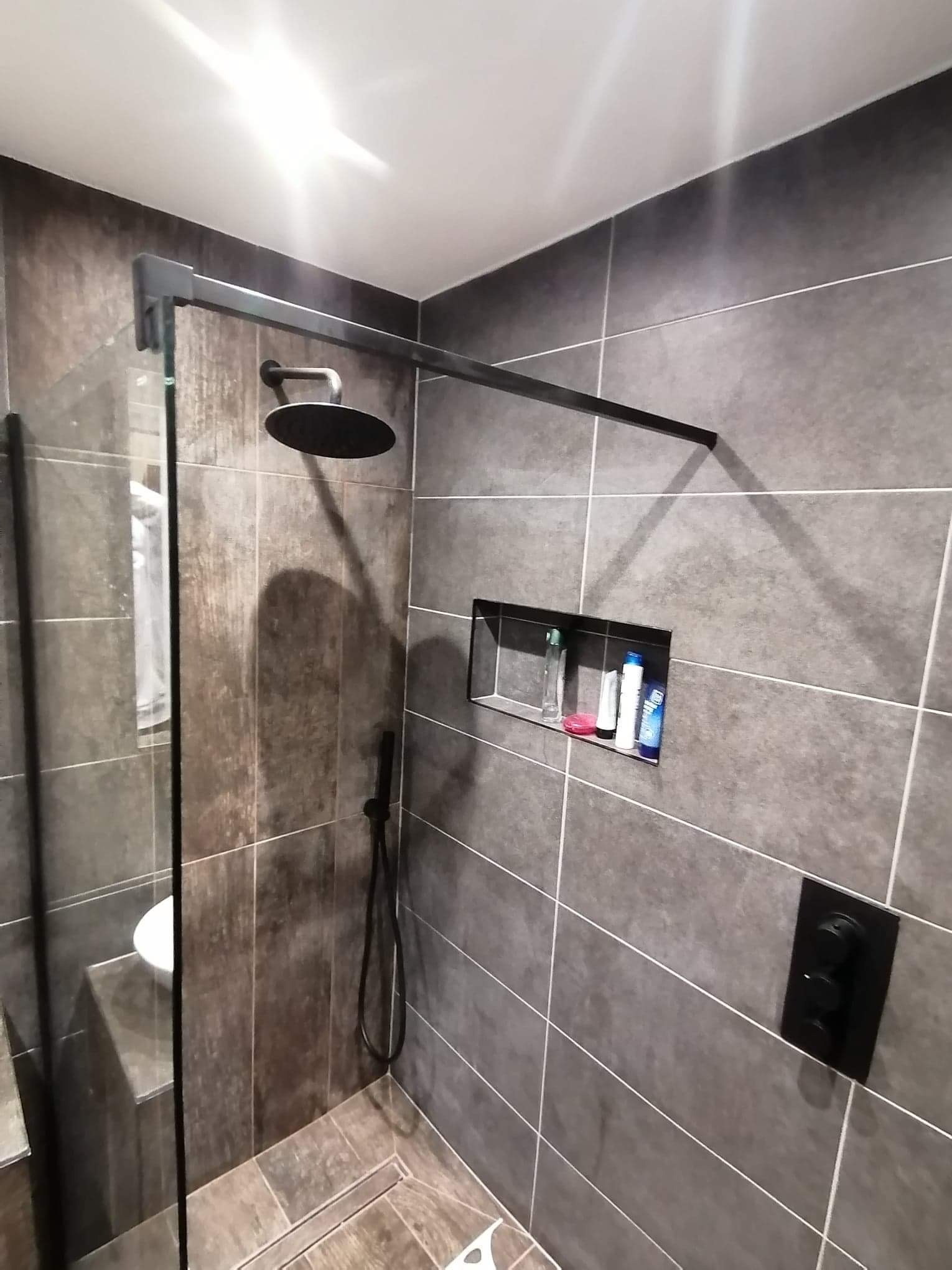 Nowoczesna kabina prysznicowa walk-in z deszczownicą i czarnymi elementami wykończeniowymi, szare płytki z wnęką na kosmetyki.