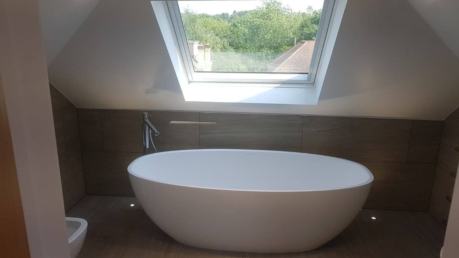 Nowoczesna łazienka na poddaszu z owalną białą wanną wolnostojącą, drewnopodobne płytki ścienne i podłogowe, okno dachowe z widokiem na zieleń.