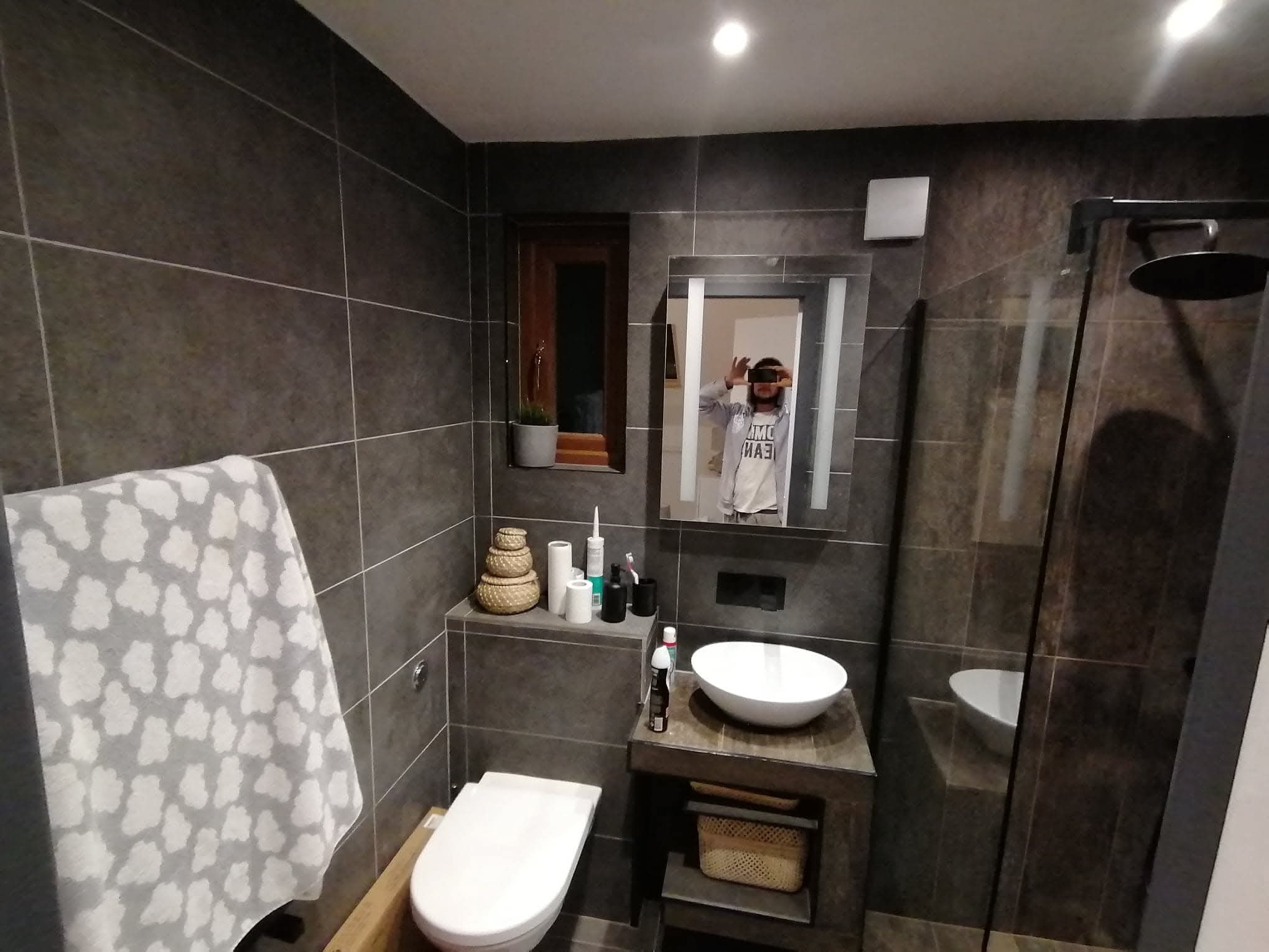 Nowoczesna łazienka z ciemnymi, grafitowymi płytkami, kabiną prysznicową typu walk-in, wiszącą miską WC i umywalką nablatową. W lustrze odbija się osoba robiąca zdjęcie.