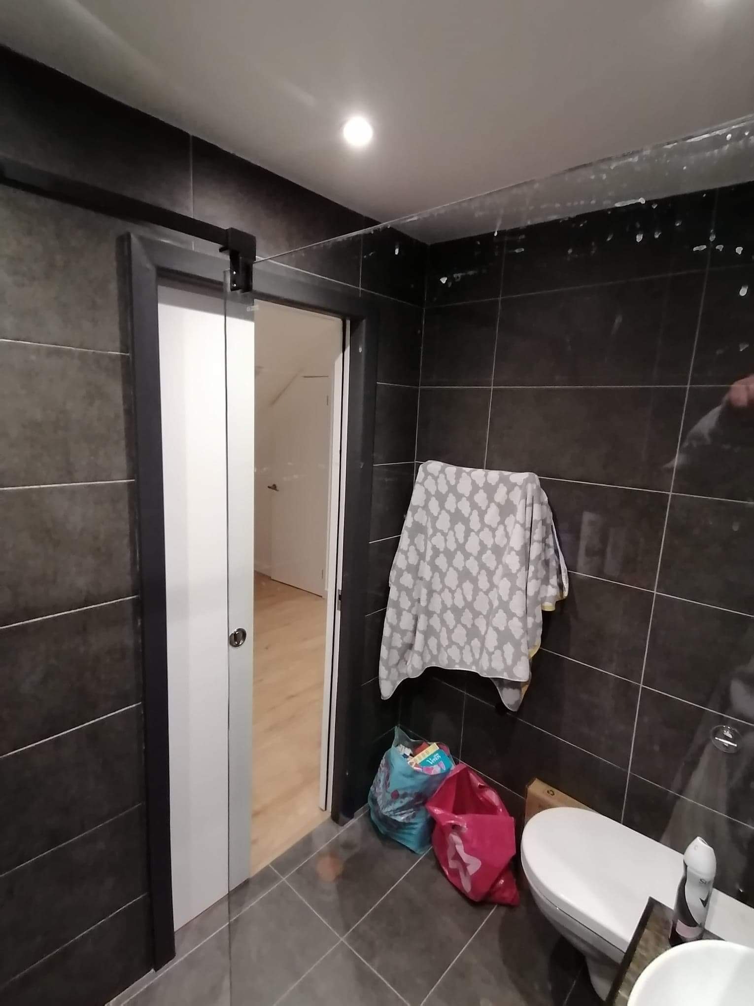 Łazienka z ciemnymi płytkami, przesuwne szklane drzwi z czarną ramą, wiszący ręcznik w białe wzory, torby i karton na podłodze, widoczne ślady po pracach wykończeniowych na suficie.