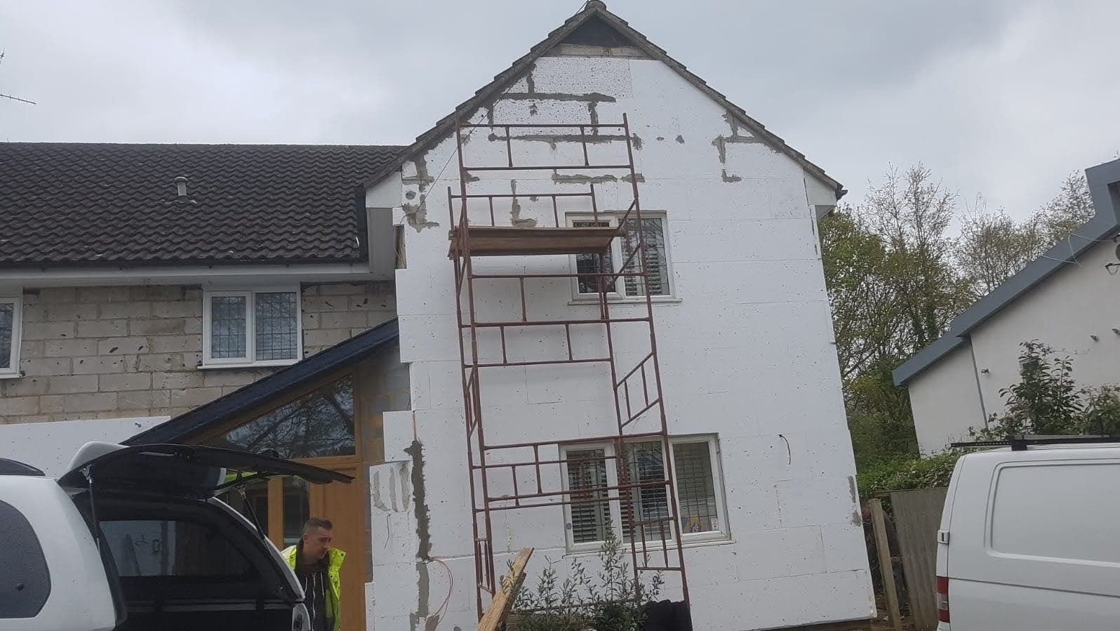Dom w trakcie ocieplania styropianem, widoczne rusztowanie, fragment samochodu z otwartą klapą bagażnika i pracownik w kamizelce odblaskowej.