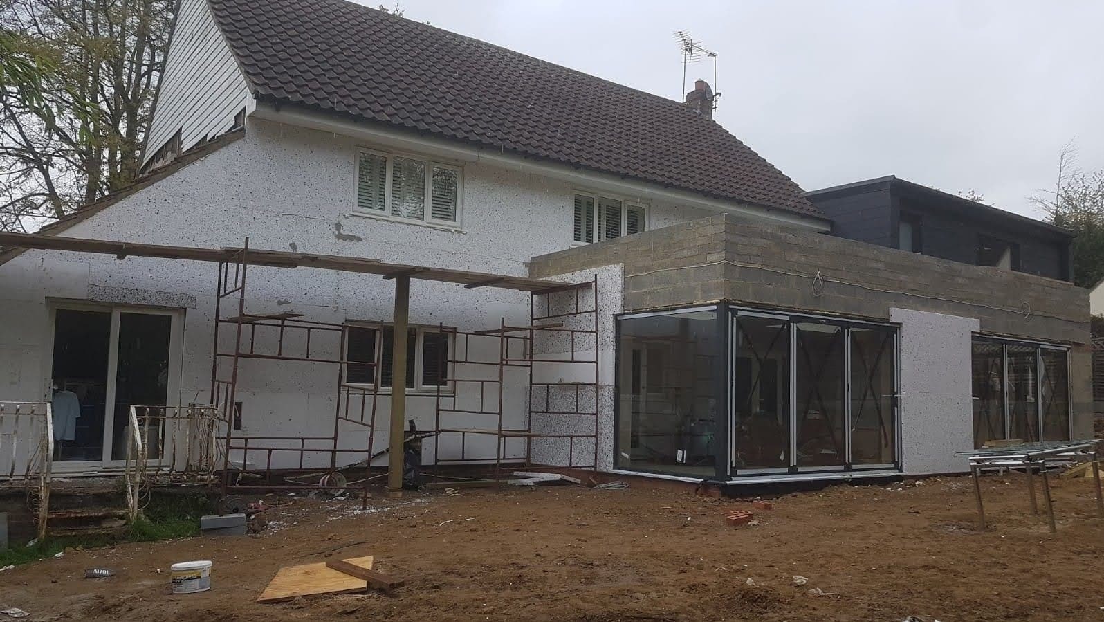 Dom jednorodzinny w trakcie rozbudowy, widoczne rusztowania i nowa część z dużymi szklanymi drzwiami przesuwnymi, ściany w trakcie ocieplania styropianem.