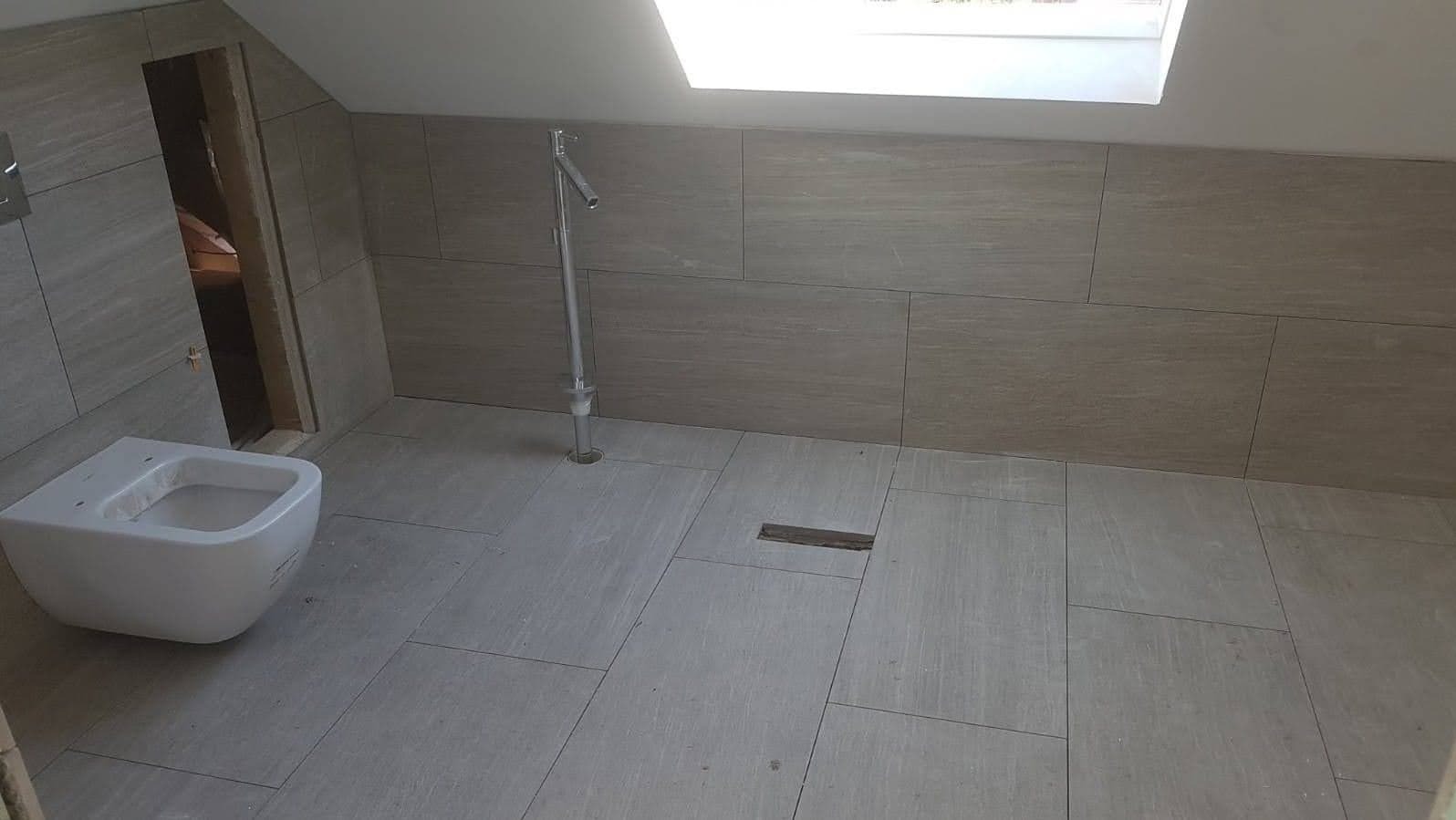 Wnętrze łazienki w trakcie prac wykończeniowych na poddaszu, widoczne płytki na ścianach i podłodze, wisząca muszla klozetowa, pionowa rura doprowadzająca wodę i otwór w podłodze, przez okno...