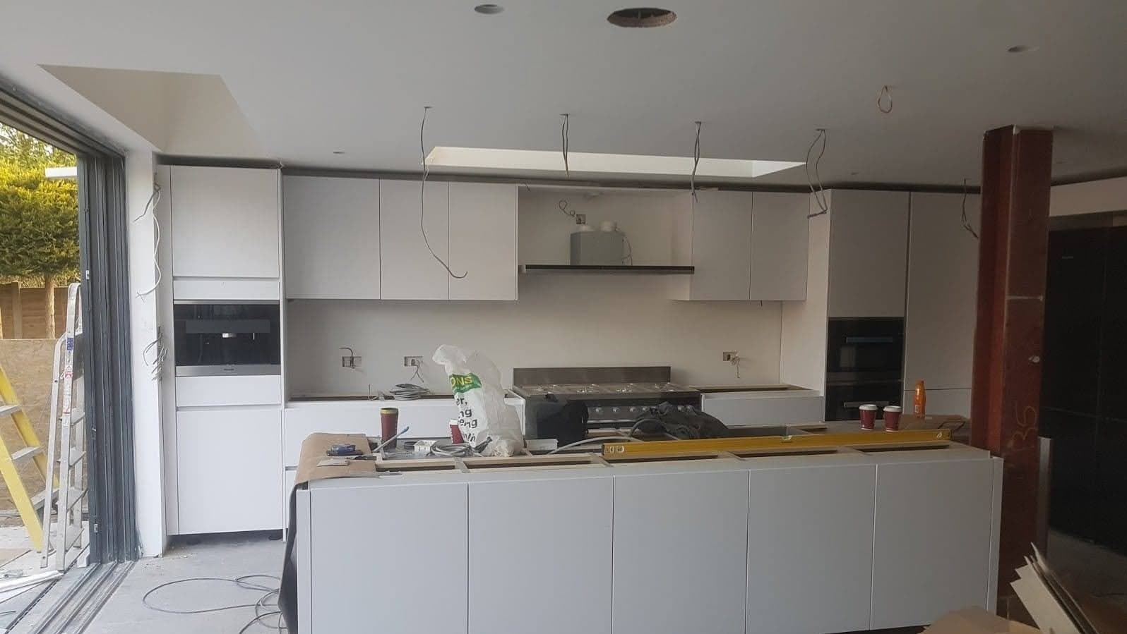 Montaż białych, minimalistycznych szafek kuchennych z widocznymi niedokończonymi instalacjami elektrycznymi i otworami w suficie pod oświetlenie, na pierwszym planie wyspa kuchenna z poziomicą.
