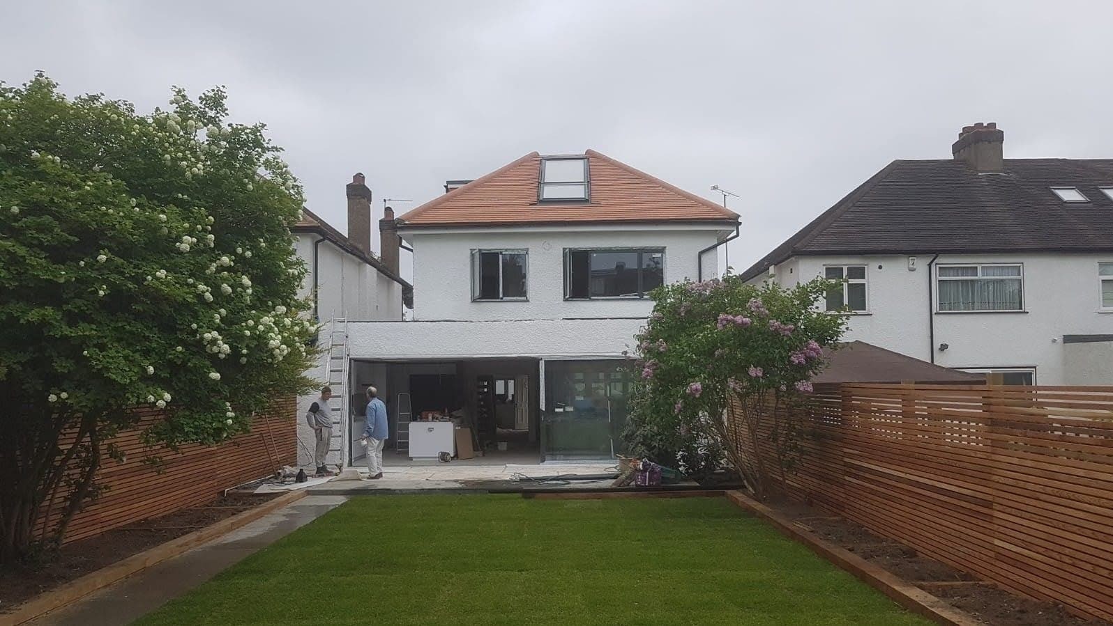 Widok z ogrodu na dom w trakcie remontu z nową elewacją, oknami i przeszkleniami, oraz drewnianym ogrodzeniem w tle, dwóch pracowników w roboczych ubraniach stoi przy ścianie domu