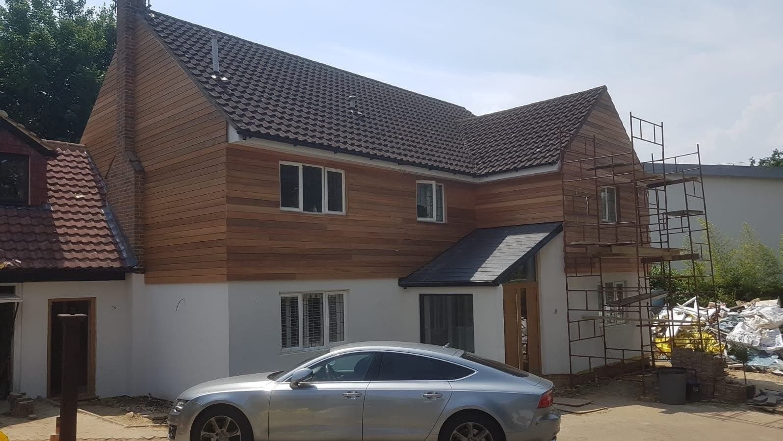 Dom jednorodzinny z nową drewnianą elewacją i rusztowaniem, częściowo otynkowany na biało, z zaparkowanym srebrnym samochodem osobowym przed budynkiem.