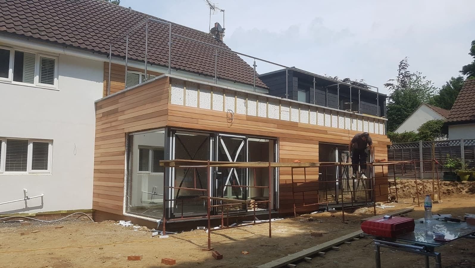Rozbudowa domu z drewnianą elewacją w trakcie montażu, widoczne okna przesuwne i pracownik na rusztowaniu.