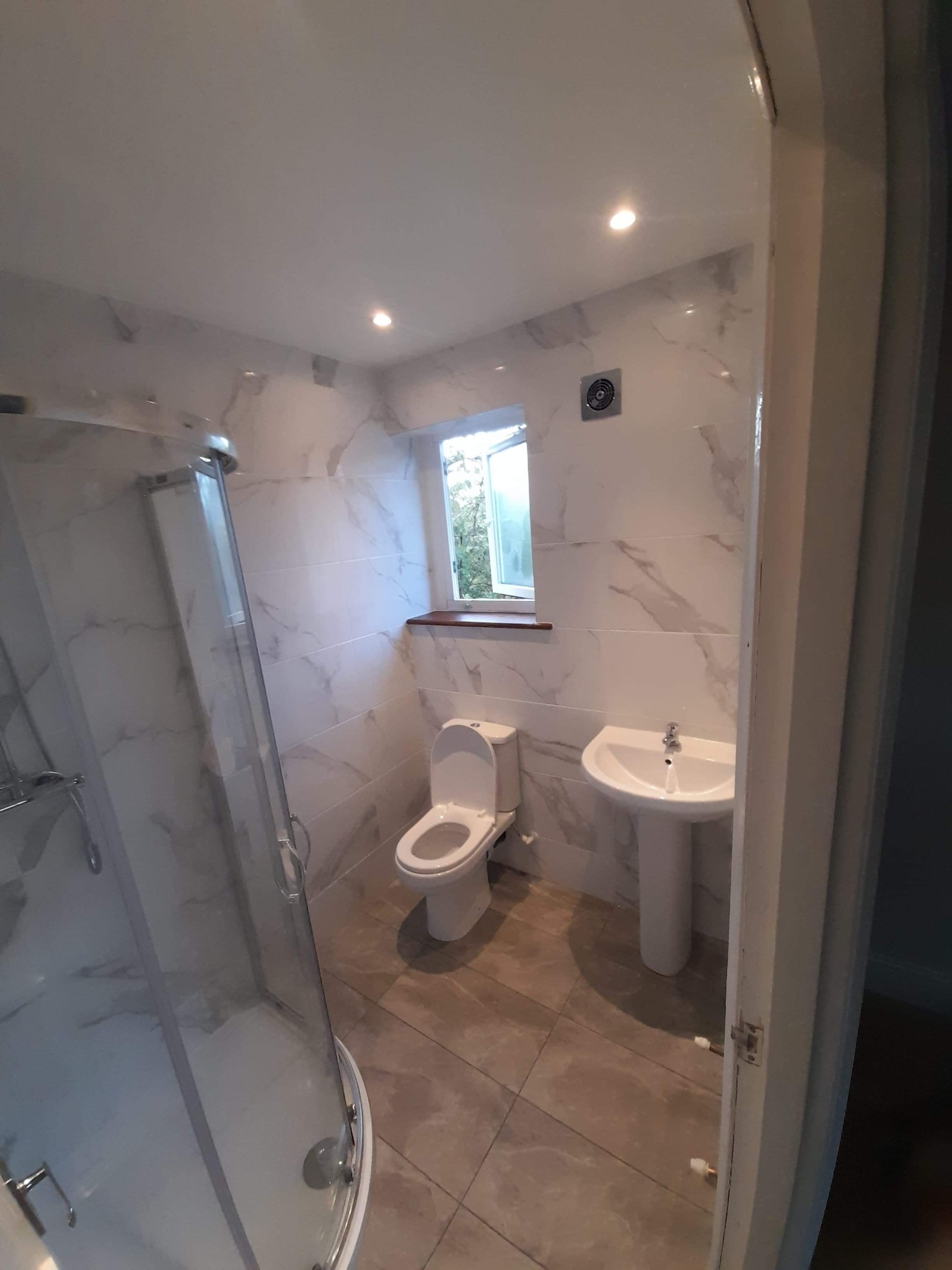 Nowoczesna łazienka z kabiną prysznicową, toaletą i umywalką na postumencie, wykończona płytkami imitującymi marmur, z oknem nad toaletą i kratką wentylacyjną na ścianie.