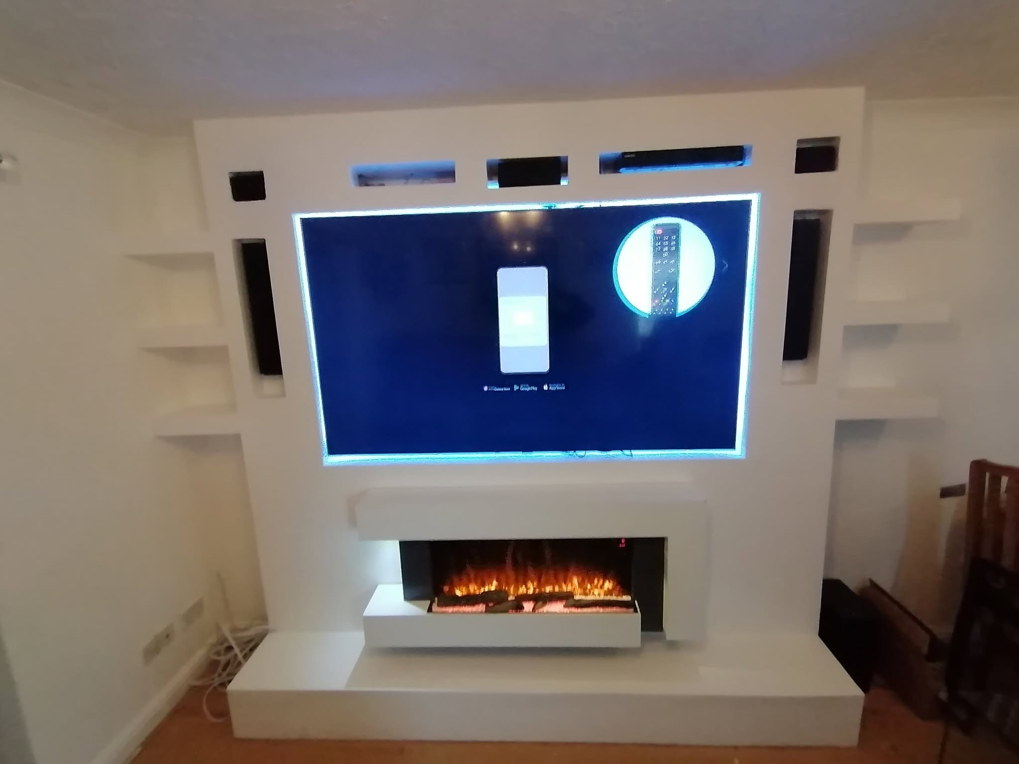 Biała zabudowa ściany z kominkiem elektrycznym i wnękami na sprzęt RTV, widoczne głośniki i półki, na ekranie telewizora ikony aplikacji.