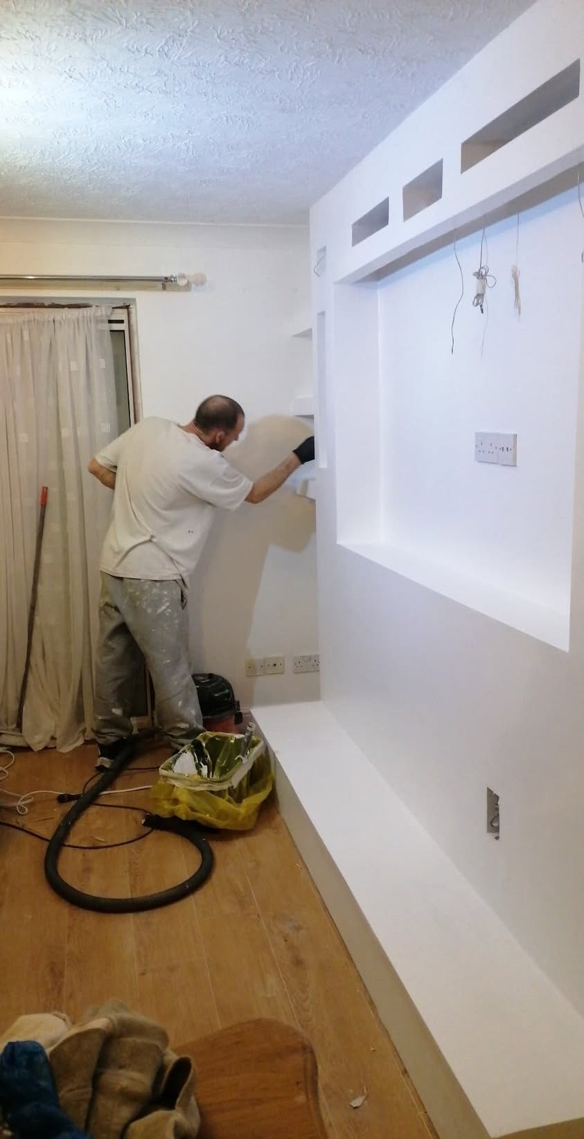 Mężczyzna w trakcie wygładzania ściany z płyty karton gipsowej, widoczna konstrukcja z wnękami, szpachla nałożona na ścianę, podłoga zabezpieczona folią, odkurzacz przemysłowy obok.