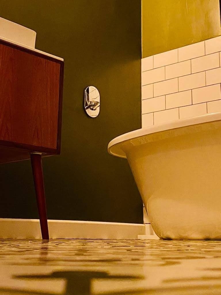 Retro łazienka z ciemnozieloną ścianą, białą wanną na nóżkach, brązową szafką pod umywalką i wzorzystą podłogą, uchwyt na ręcznik na ścianie