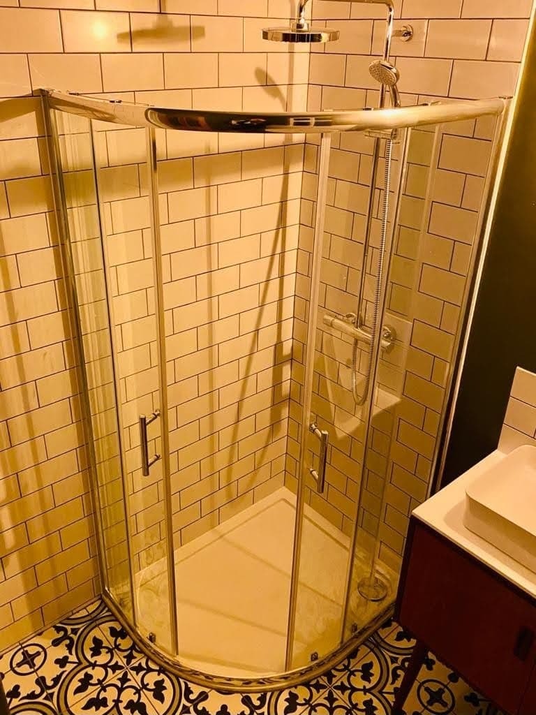 Narożna kabina prysznicowa z chromowanymi profilami i uchwytami, wyłożona płytkami imitującymi cegłę w ciepłym odcieniu, z dekoracyjną podłogą w geometryczny wzór.