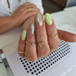 Dłoń z owalnymi paznokciami, jeden w odcieniu pistacjowej zieleni, drugi pokryty brokatem, ozdobiona złotymi pierścionkami na tle lampy UV do utwardzania lakieru hybrydowego.