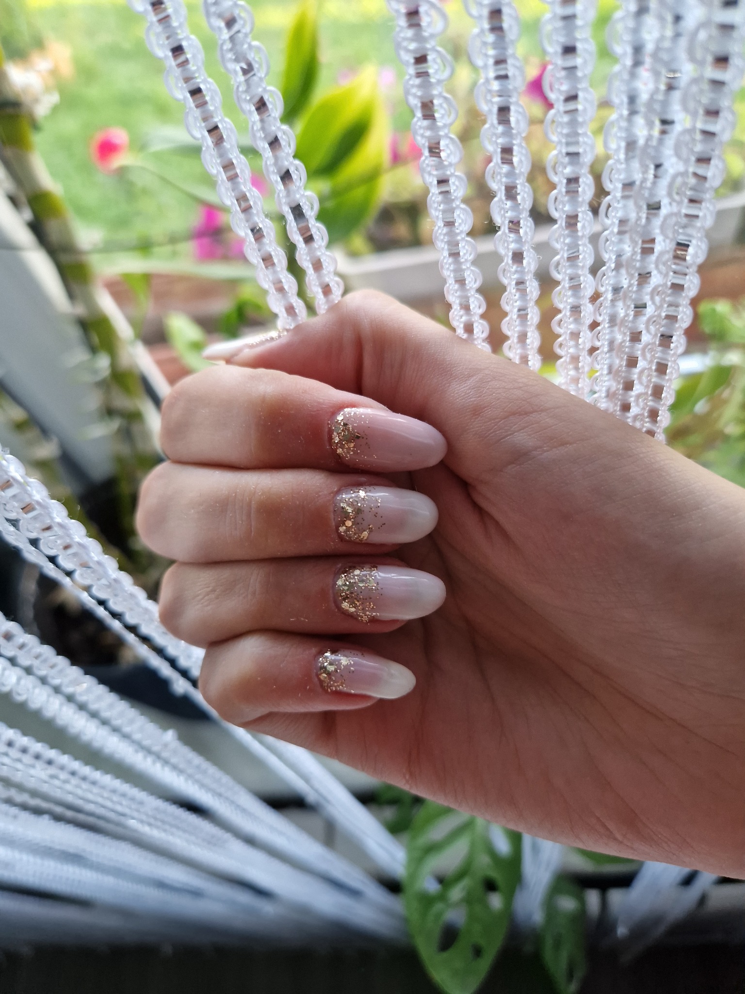 Dłoń z owalnymi paznokciami w kolorze nude, ozdobionymi złotym brokatem na końcach, na tle białej, koralikowej firanki i zieleni roślin.