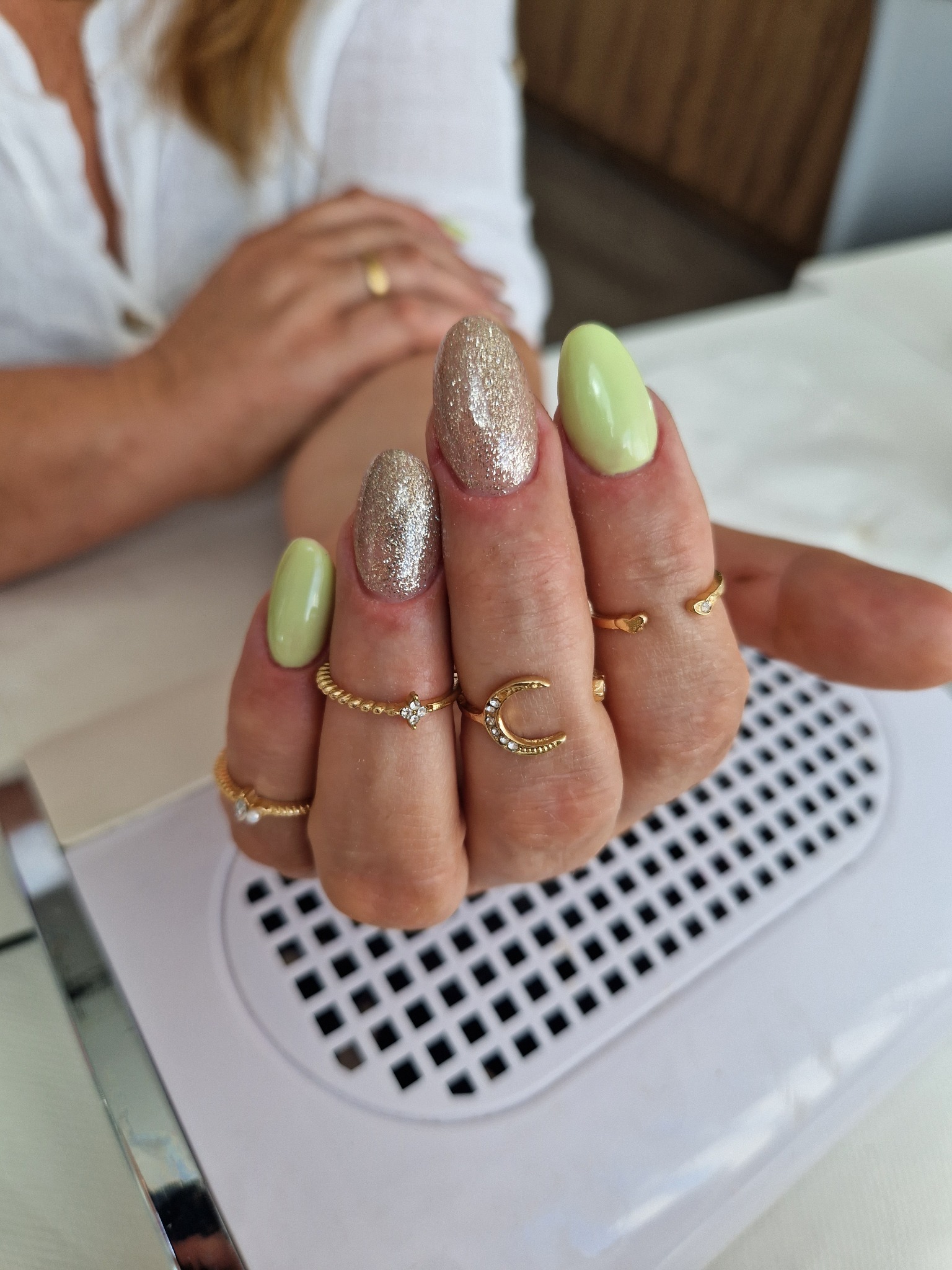 Dłoń z owalnymi paznokciami, jeden w odcieniu pistacjowej zieleni, drugi pokryty brokatem, ozdobiona złotymi pierścionkami na tle lampy UV do utwardzania lakieru hybrydowego.