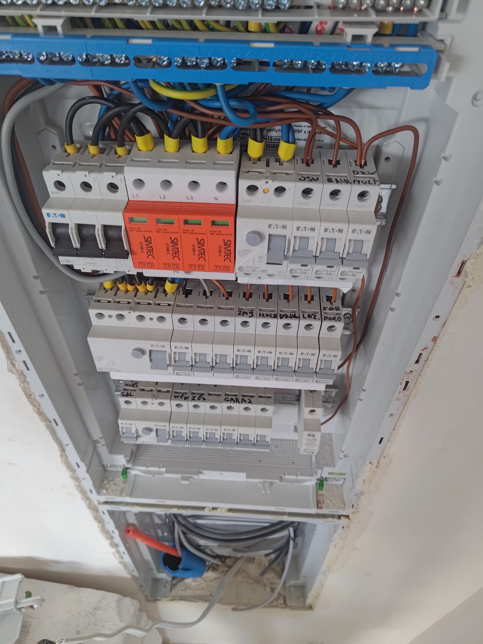 Rozdzielnica elektryczna z zabezpieczeniami przepięciowymi, bezpiecznikami i przewodami oznaczonymi żółtymi końcówkami, zamontowana w ścianie z widocznymi śladami prac budowlanych.