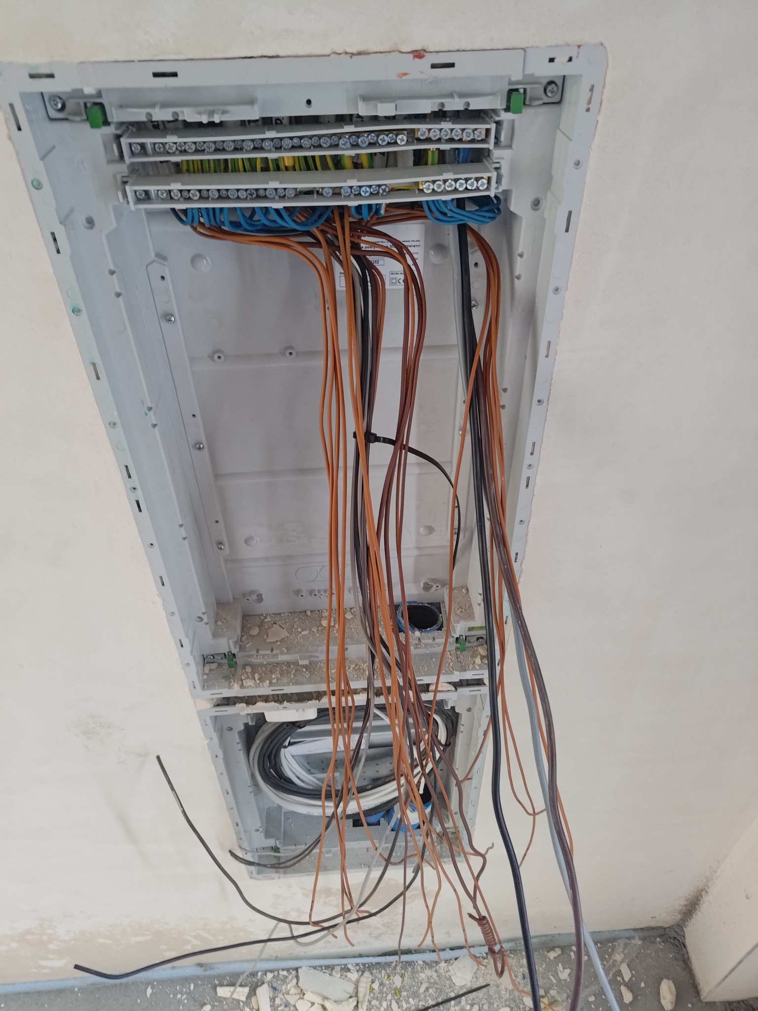 Otwarta skrzynka rozdzielcza na ścianie z widocznymi przewodami elektrycznymi w różnych kolorach, w trakcie montażu, z fragmentami gruzu na podłodze.
