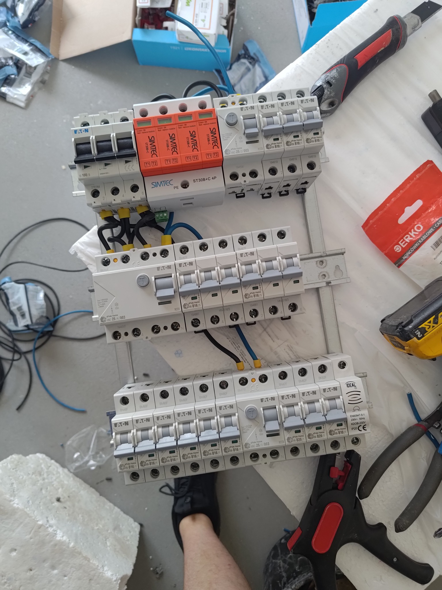 Rozdzielnica elektryczna w trakcie montażu, widoczne zabezpieczenia przepięciowe Simtec, wyłączniki Eaton, przewody, narzędzia: nóż monterski, obcęgi do kabli, zaciskarka do końcówek,...