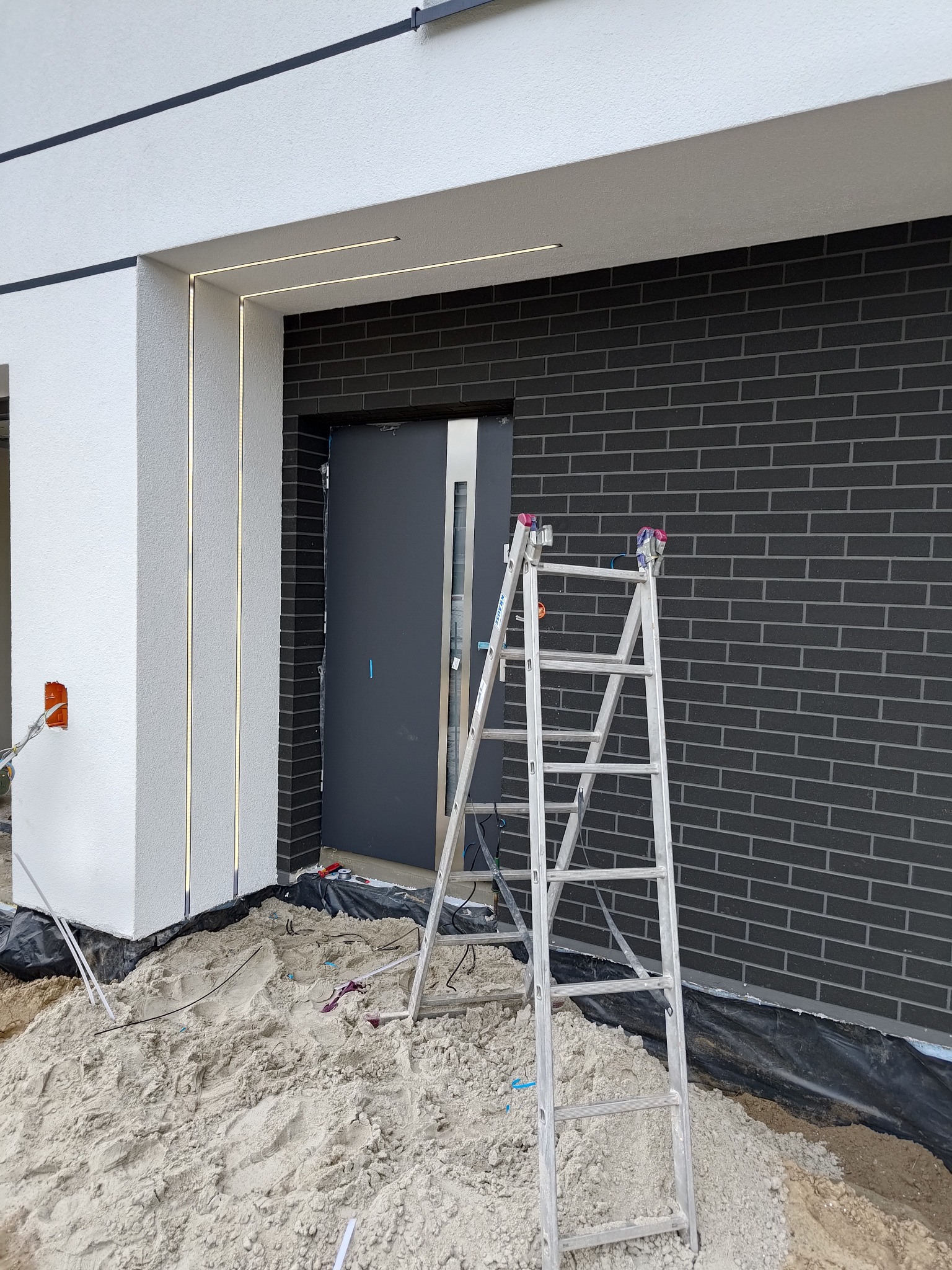 Nowoczesny dom w trakcie montażu oświetlenia zewnętrznego; widoczna aluminiowa drabina oparta o elewację z cegły klinkierowej, świecące linearne oprawy wkomponowane w elewację, piasek na podłożu.