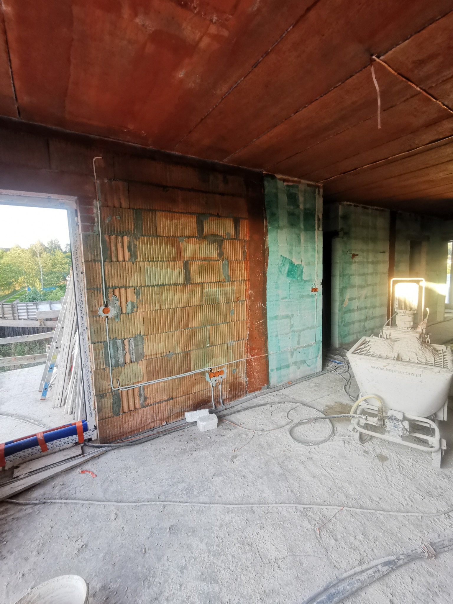 Wnętrze w trakcie prac instalacyjnych: ściany z pustaków, widoczne przewody elektryczne w peszlach, puszki instalacyjne, agregat tynkarski Bapro na zewnątrz budynku.