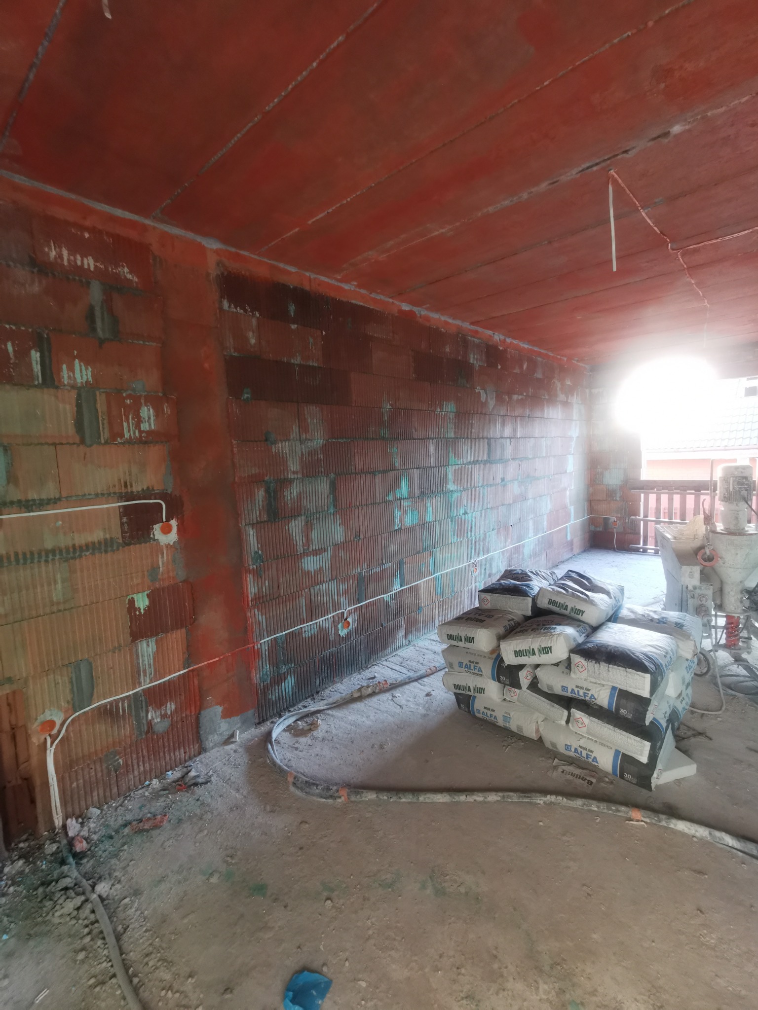 Wnętrze w trakcie prac wykończeniowych: widoczne pakiety zaprawy Dolina Nidy, ściany z pustaków, instalacja elektryczna w peszlach i maszyna do nakładania tynku.