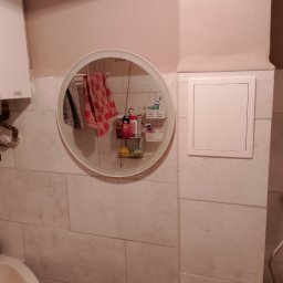 Krzysztof Ścibura - Łazienka z beżowymi płytkami, okrągłe lustro odbija półki z kosmetykami i ręcznikami, widoczny fragment umywalki.