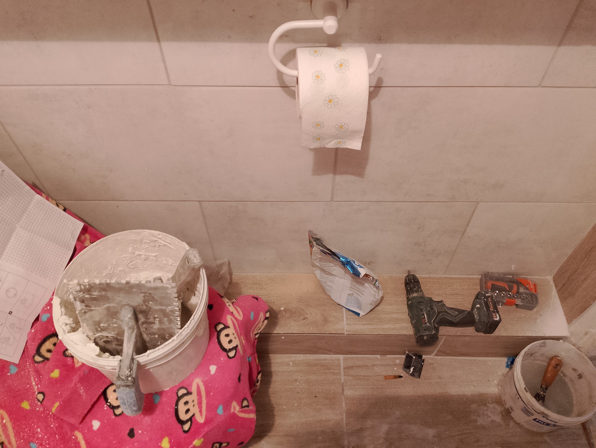 Wiadro z zaprawą szpachlową i kielnią na różowej bluzie dziecięcej, w tle płytki ceramiczne, wiertarka akumulatorowa i opakowanie po produkcie budowlanym.