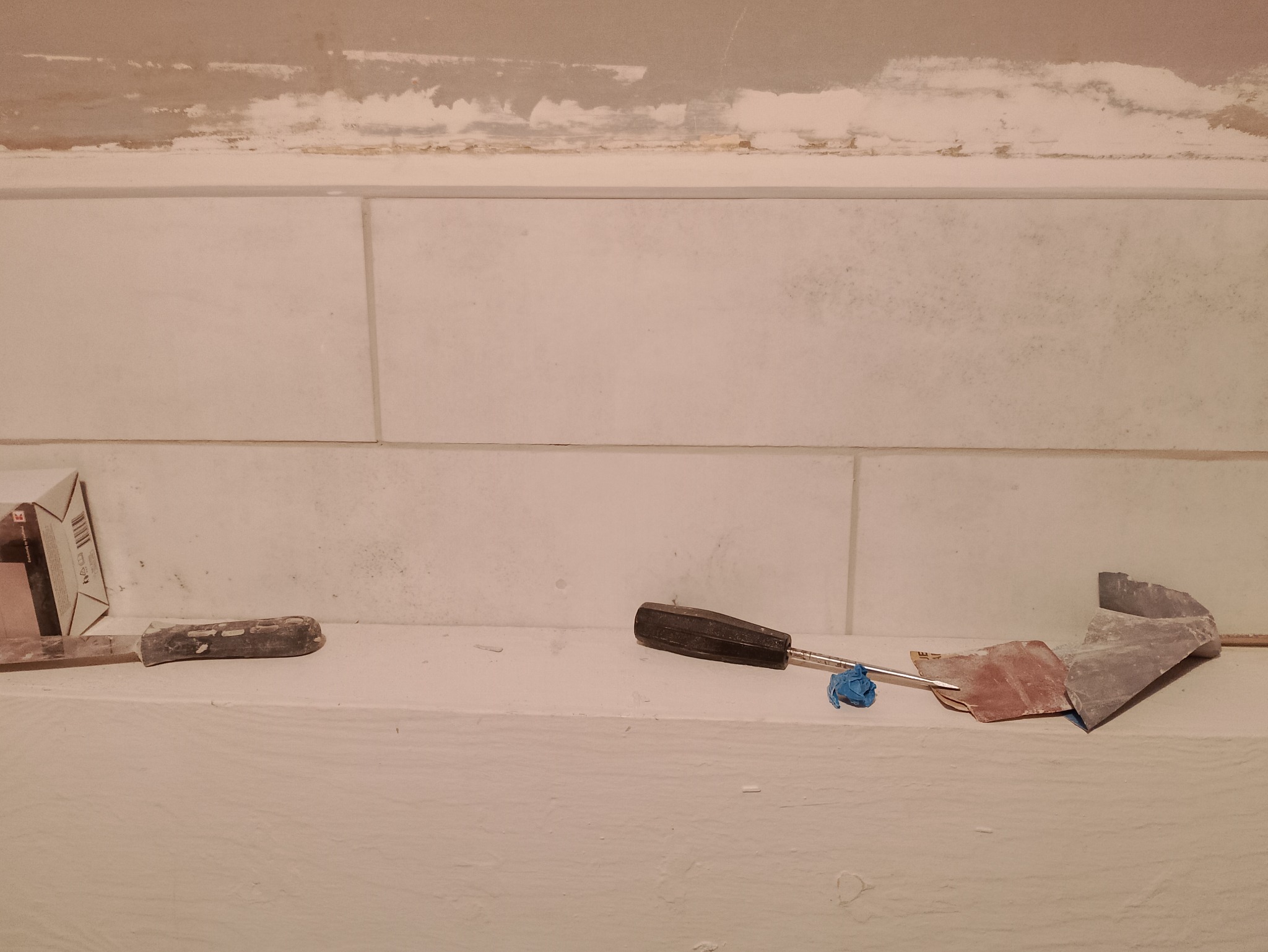 Ściana z jasnych, prostokątnych płyt, z widocznymi narzędziami: szpachelka z czarną rączką, pacą do szpachlowania i opakowaniem po kleju do płytek, wskazującymi na etap prac wykończeniowych.