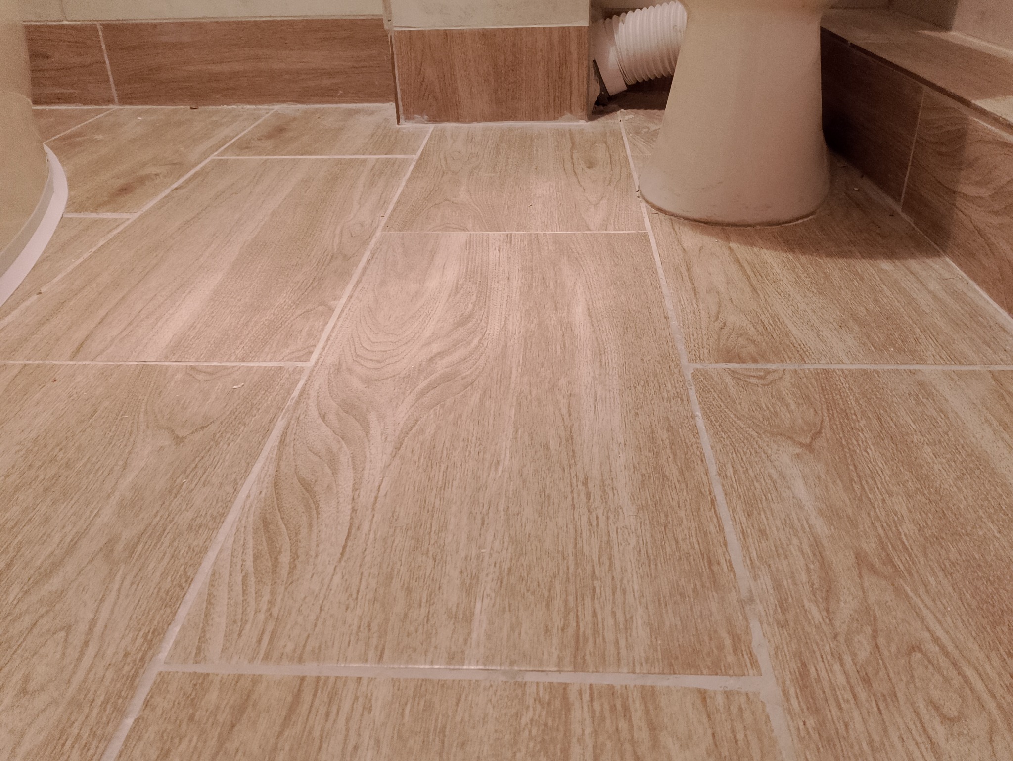 Ułożone płytki podłogowe imitujące drewno w łazience, widoczne fragment toalety i dolna część ściany z podobnymi płytkami.