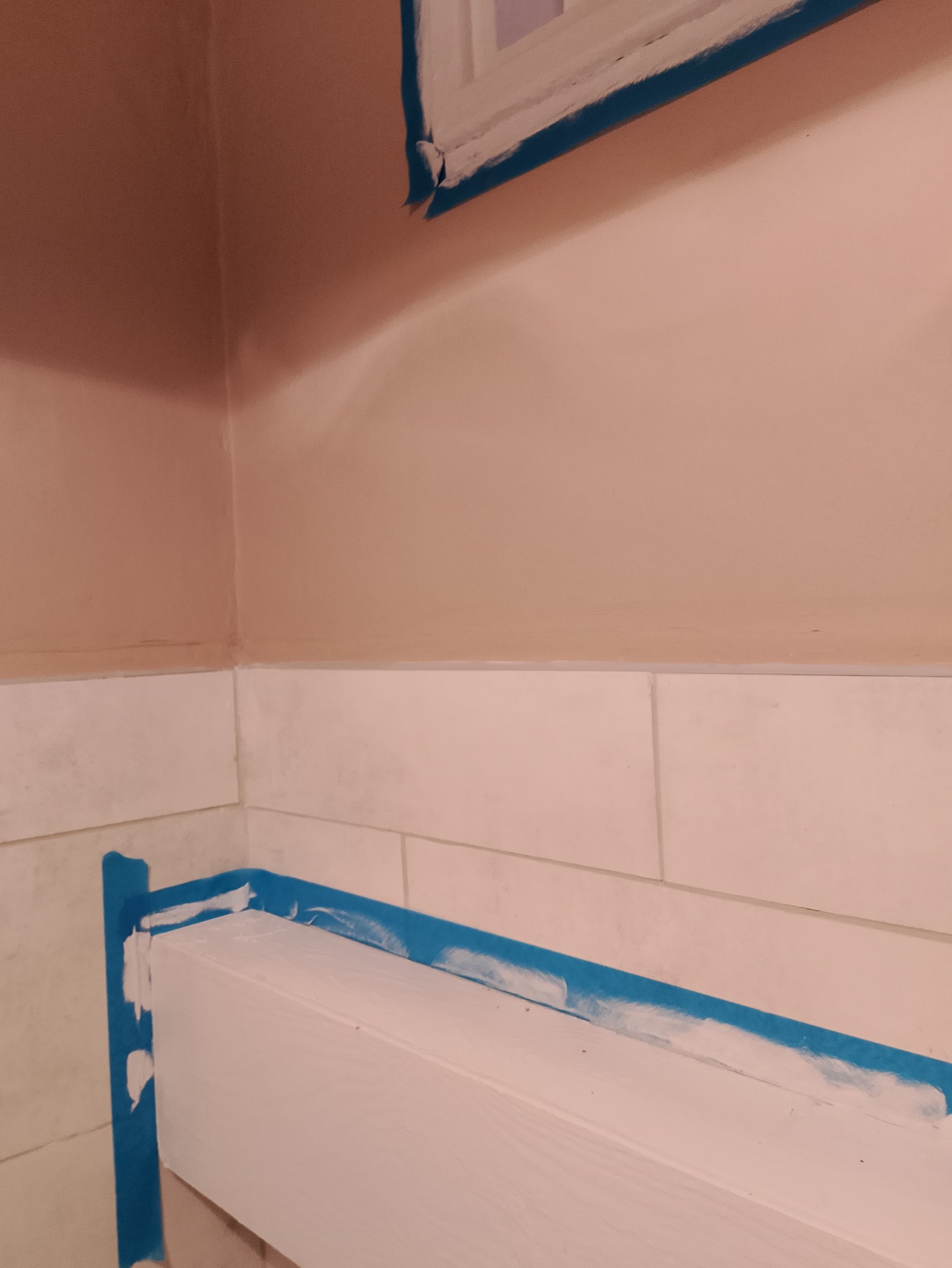 Ściana i listwa przypodłogowa zabezpieczone taśmą malarską, fragment okna w górnym rogu, ślady świeżego malowania.