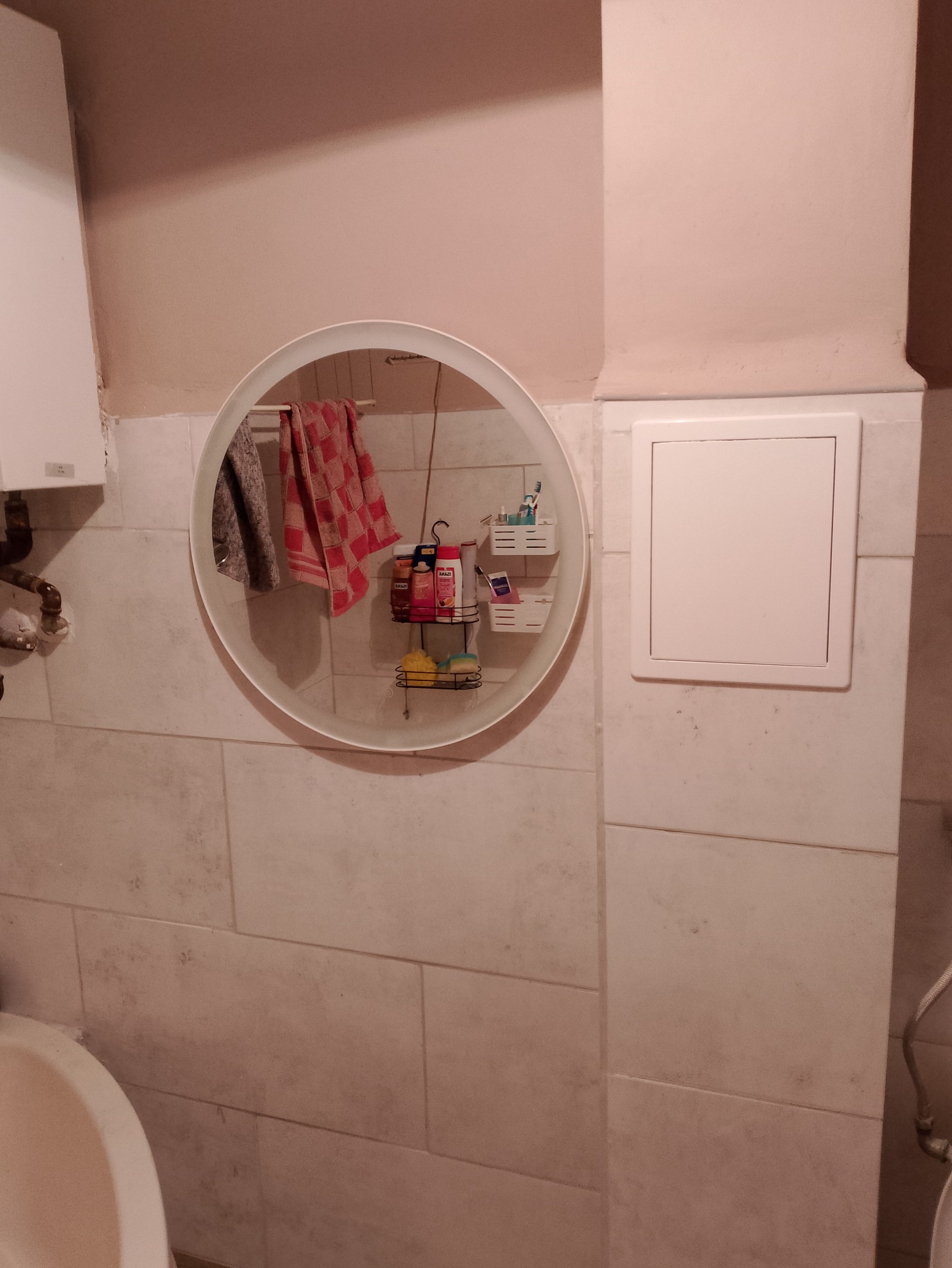 Łazienka z beżowymi płytkami, okrągłe lustro odbija półki z kosmetykami i ręcznikami, widoczny fragment umywalki.