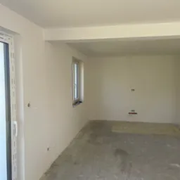 Wnętrze w stanie deweloperskim z oknami PCV, świeżo wygładzonymi ścianami i surowym betonem na podłodze, przygotowane do dalszych prac wykończeniowych.