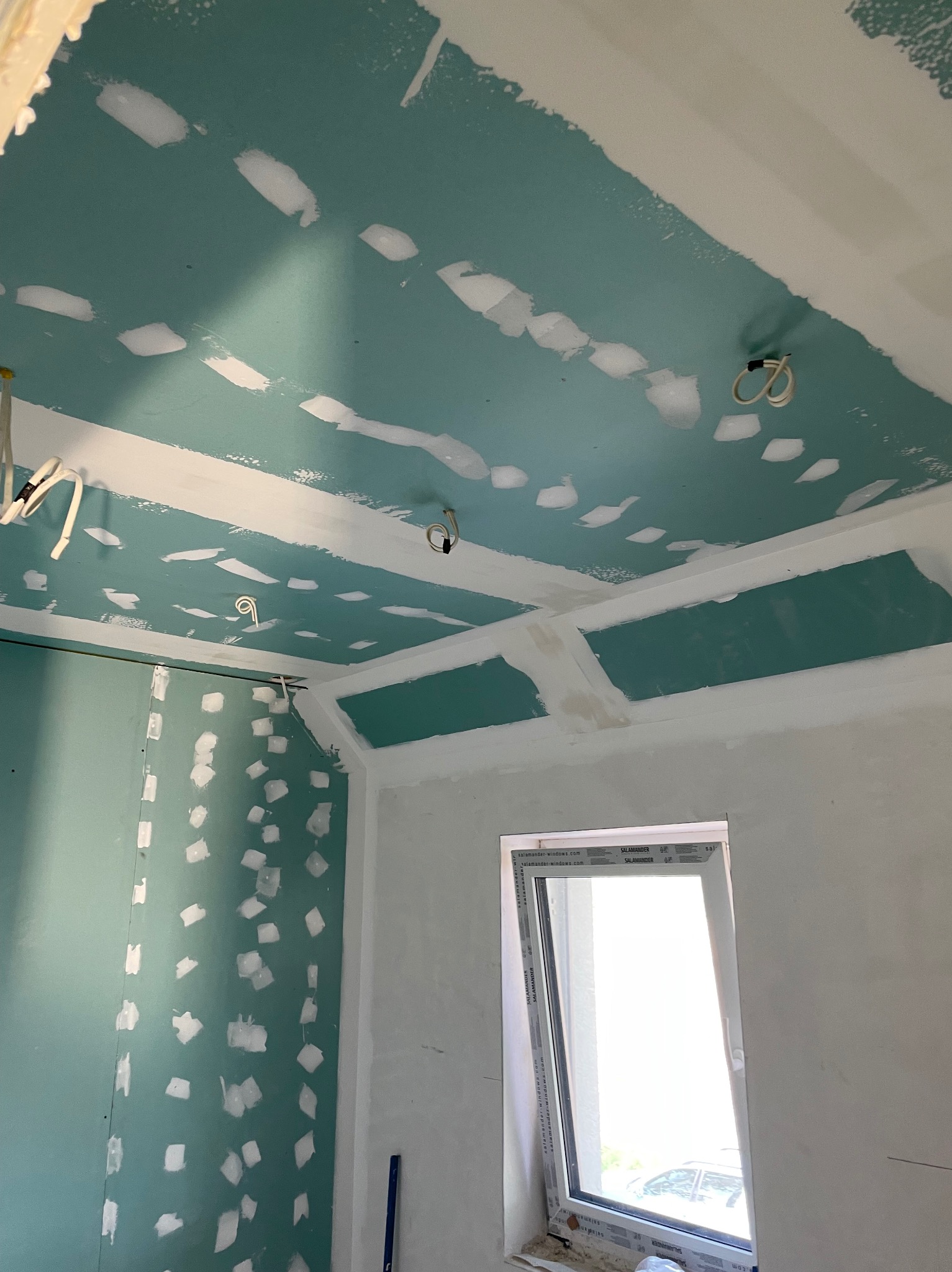 Ściana i sufit z płyt kartonowo-gipsowych, wstępnie przygotowane do dalszych prac wykończeniowych, z widocznymi punktowymi łączeniami szpachlowymi oraz wystającymi przewodami elektrycznymi, okno...